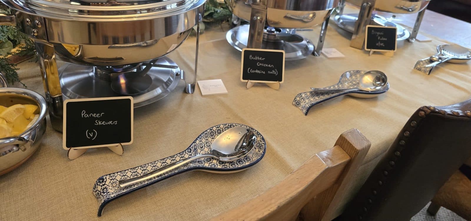 A buffet table laid with chafing dishes and patterned spoon rests, with chalkboard signs for Paneer Skewers, Butter Chicken and Biryani Pulao