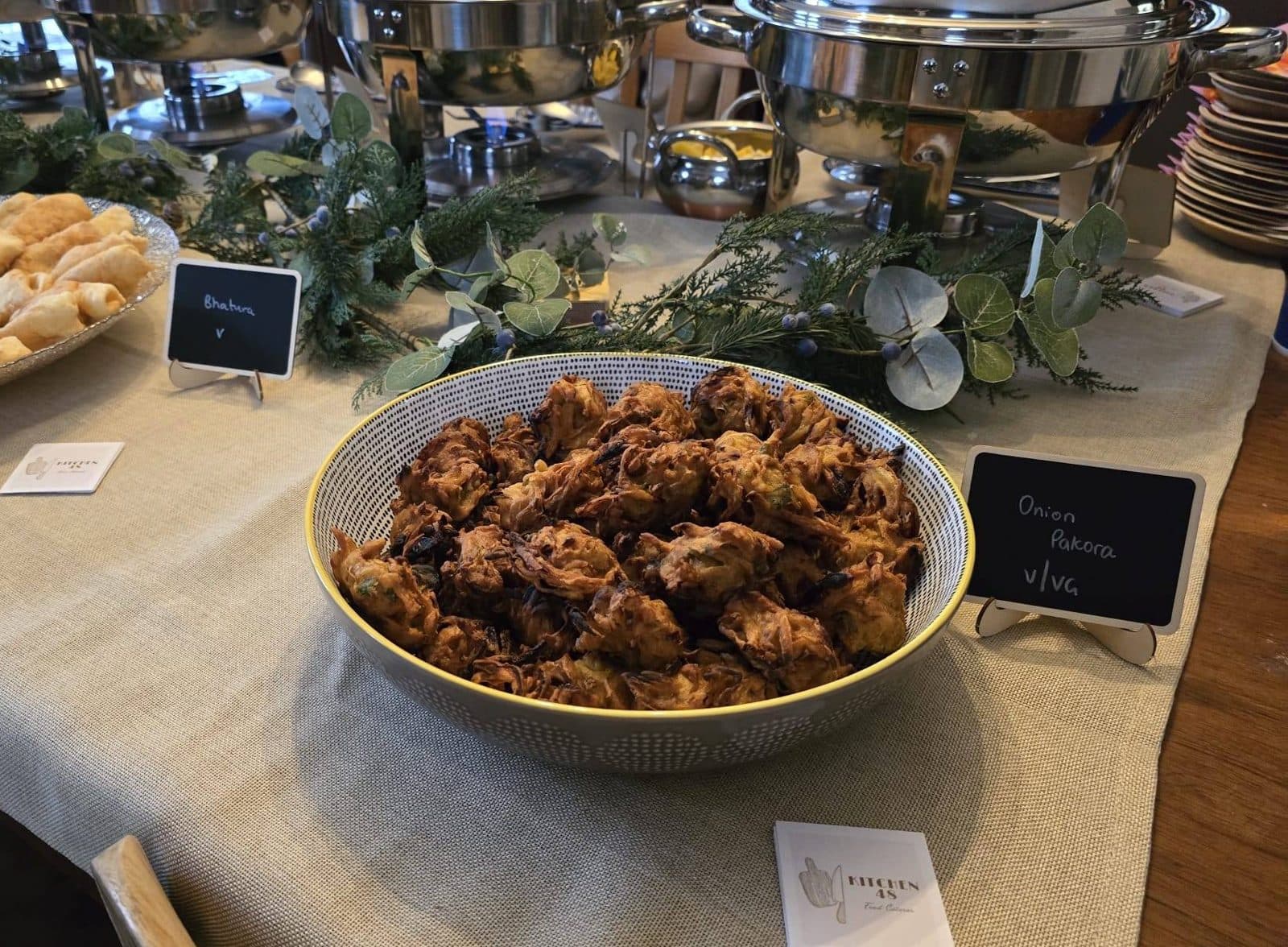 A speckled bowl piled with crisp onion pakora on a buffet table, next to a small chalkboard sign reading Onion Pakora, with bhatura and chafing dishes behind