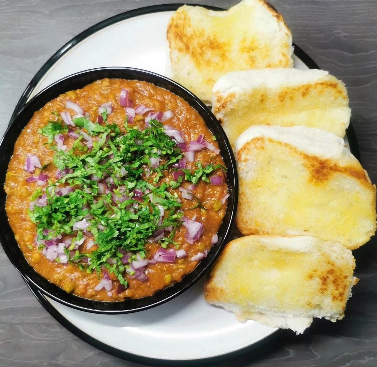 A bowl of pav bhaji topped with chopped red onion and fresh coriander, served with four slices of toasted, buttered pav