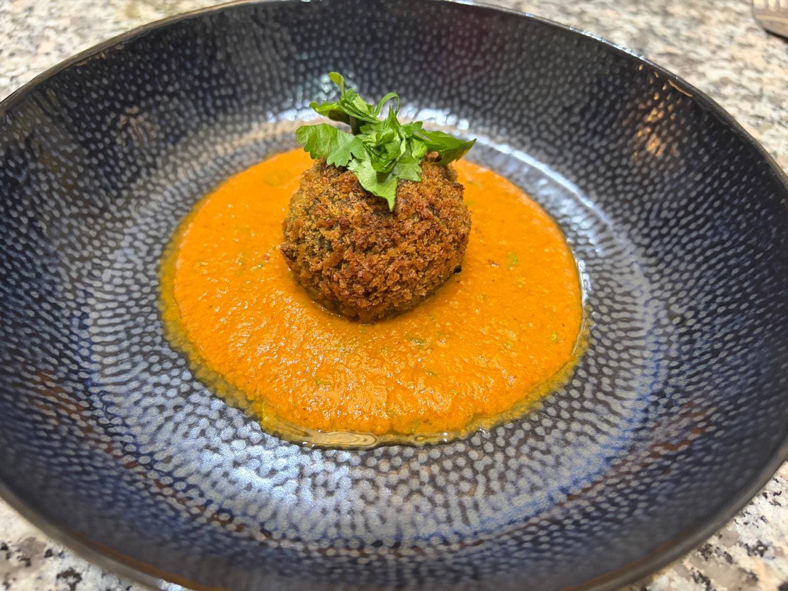 A golden, crumb-coated arancini topped with fresh green herbs, sitting in a pool of bright orange makhani sauce in a dark speckled bowl